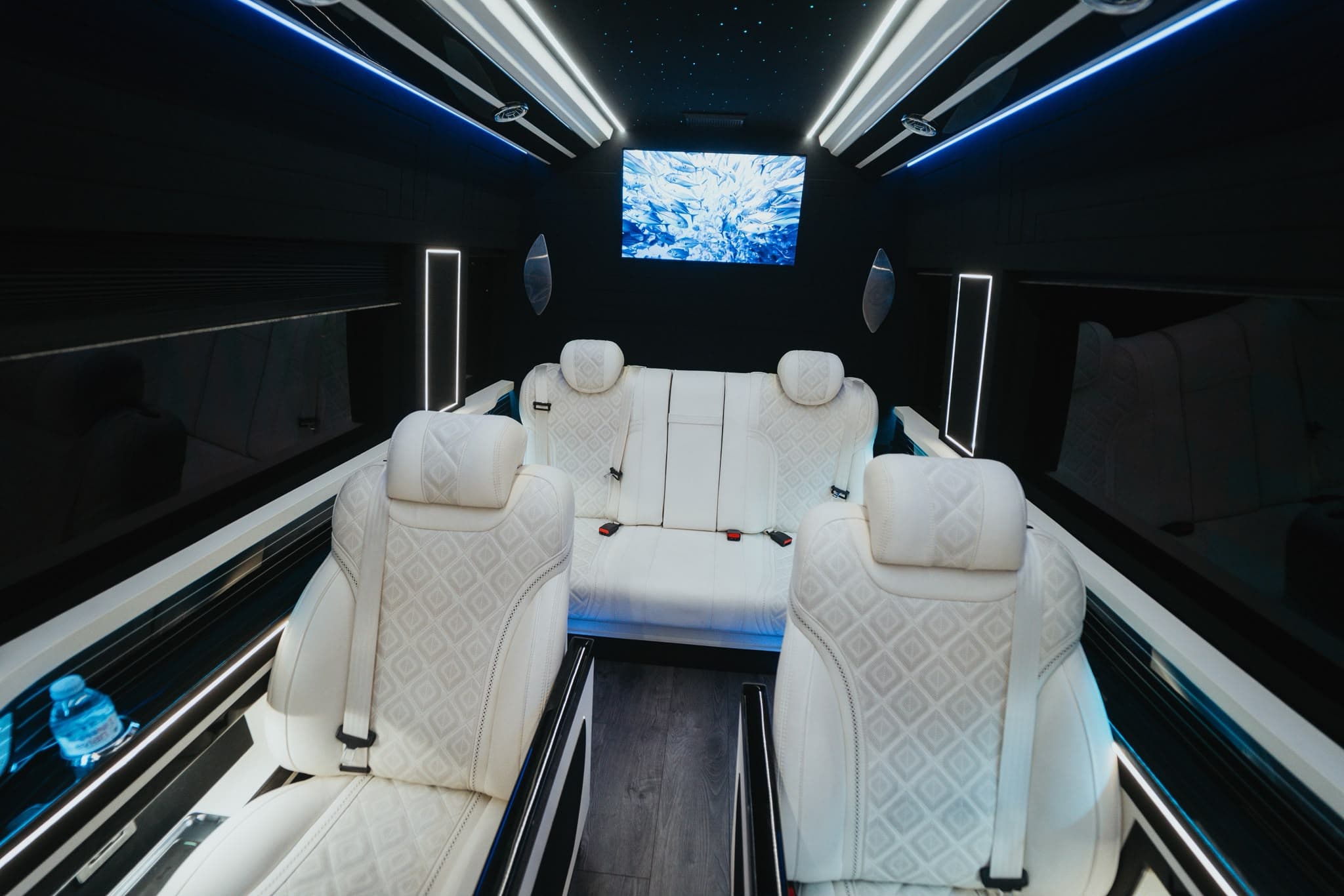 Intérieur du Sprinter VIP Starlight avec ciel étoilé, fauteuils capitonnés blancs face-à-face et écran géant